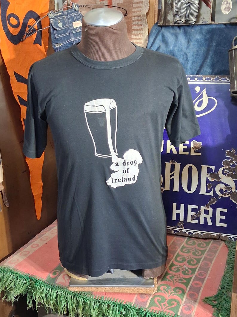 Op de afbeelding: Zwart t-shirt met een witte afbeelding van een pint bier en de omtrek van Ierland, met de tekst "a drop of Ireland". Het t-shirt wordt op een mannequin gepresenteerd.