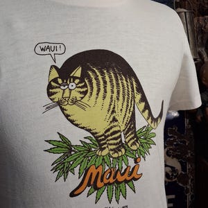 May include: White t-shirt featuring a cartoon cat with brown and yellow stripes, perched on green leaves. The word "Maui" is in orange script below the cat, with the text "WAUI!" in a speech bubble. The artist's signature and the year 1975 are at the bottom.