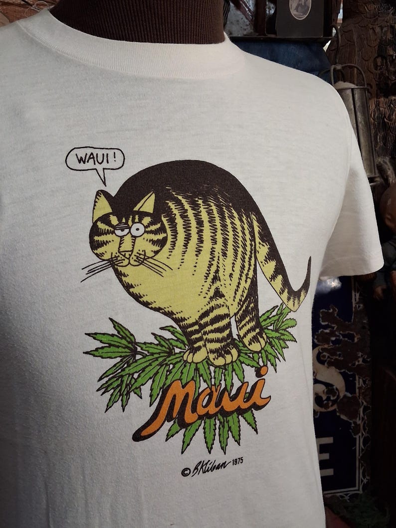 May include: White t-shirt featuring a cartoon cat with brown and yellow stripes, perched on green leaves. The word "Maui" is in orange script below the cat, with the text "WAUI!" in a speech bubble. The artist's signature and the year 1975 are at the bottom.