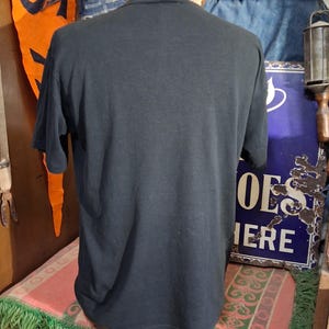 May include: A black short-sleeved t-shirt displayed on a mannequin. The shirt is plain and appears to be made of a soft material. The background includes vintage decor and signage.