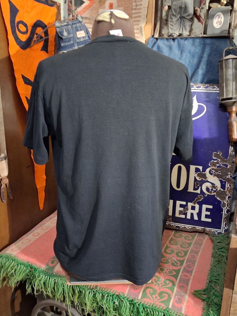 May include: A black short-sleeved t-shirt displayed on a mannequin. The shirt is plain and appears to be made of a soft material. The background includes vintage decor and signage.