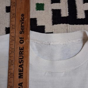 K&ouml;nnte beinhalten: Nahaufnahme eines wei&szlig;en T-Shirts mit Rundhalsausschnitt. Ein Holzlineal mit dem Text "EXTRA MEASURE OF SERVICE" wird vertikal platziert und zeigt Messungen in Zentimetern an.