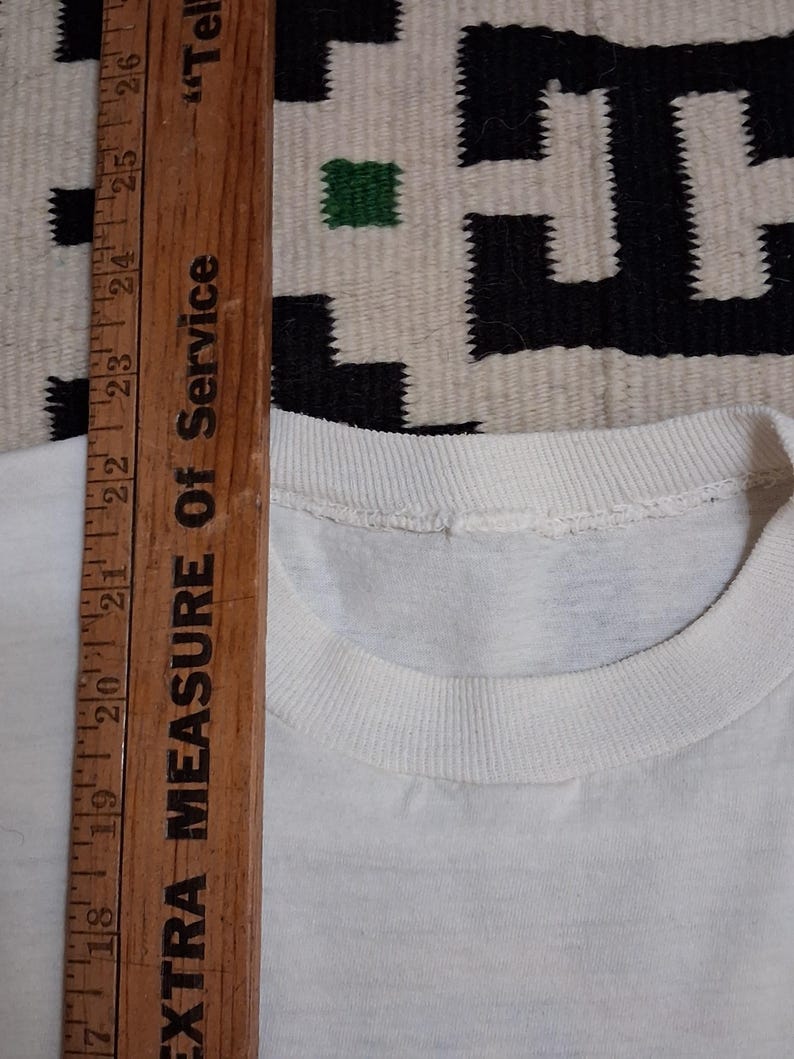 K&ouml;nnte beinhalten: Nahaufnahme eines wei&szlig;en T-Shirts mit Rundhalsausschnitt. Ein Holzlineal mit dem Text "EXTRA MEASURE OF SERVICE" wird vertikal platziert und zeigt Messungen in Zentimetern an.