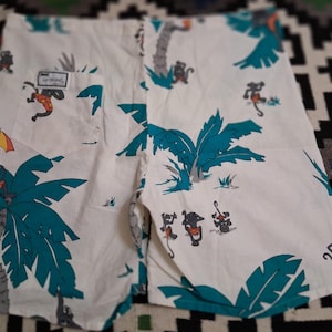 May include: White shorts with a tropical print featuring teal palm trees and cartoon monkeys. The shorts have a pocket with the text "UP FRONT". The design includes monkeys with musical instruments and an umbrella.