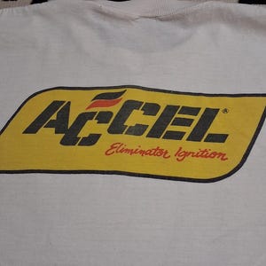 K&ouml;nnte beinhalten: Wei&szlig;es T-Shirt mit einem gelb-schwarzen ACCEL-Logo und dem roten Text "Eliminator Ignition". Das Logo hat eine rechteckige Form mit abgerundeten Ecken. Der Kragen des T-Shirts ist oben sichtbar.