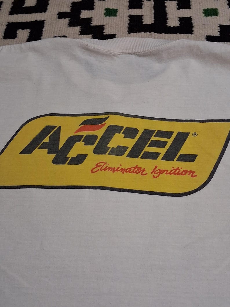 K&ouml;nnte beinhalten: Wei&szlig;es T-Shirt mit einem gelb-schwarzen ACCEL-Logo und dem roten Text "Eliminator Ignition". Das Logo hat eine rechteckige Form mit abgerundeten Ecken. Der Kragen des T-Shirts ist oben sichtbar.