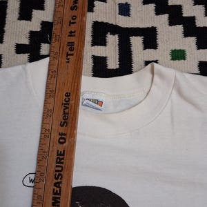 May include: Cream-colored t-shirt with a graphic illustration of a person's face and the word "W" in a speech bubble. A wooden ruler with inch measurements and the text "Tell It To Sweeney" is in the image.
