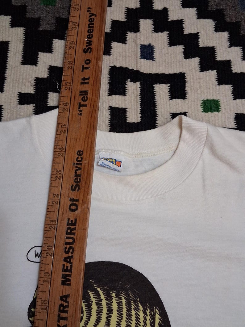 May include: Cream-colored t-shirt with a graphic illustration of a person's face and the word "W" in a speech bubble. A wooden ruler with inch measurements and the text "Tell It To Sweeney" is in the image.