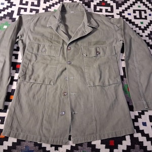 Può includere: Giacca verde oliva in stile militare con quattro tasche frontali e chiusure con bottoni. La giacca a maniche lunghe ha un collo e una vestibilità su misura. La giacca è stesa su una superficie a motivi.