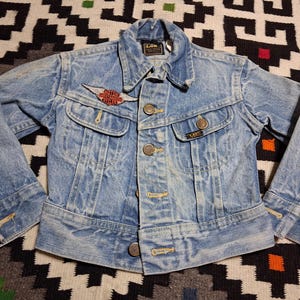 May include: A light blue denim jacket with a pointed collar, long sleeves, and metal button closures. The jacket features two chest pockets with button flaps, a Lee brand label, and a red and white winged motorcycle patch on the chest.