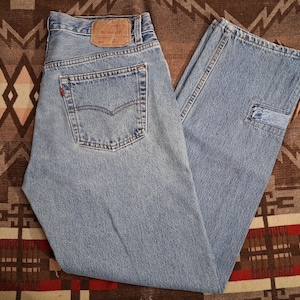 May include: Light blue denim jeans featuring a classic five-pocket design, a tan leather patch on the back waistband, a small red tab on the rear pocket, and a straight-leg cut with visible stitching details throughout the garment.