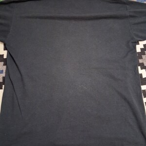 May include: A solid black t-shirt, viewed from the back. The shirt is made of a soft material and has a crew neck. The hem and sleeves are visible. The shirt is laying on a patterned surface.
