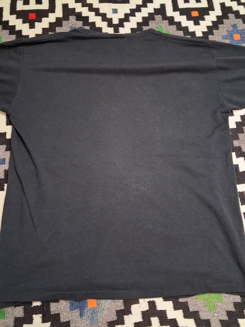 May include: A solid black t-shirt, viewed from the back. The shirt is made of a soft material and has a crew neck. The hem and sleeves are visible. The shirt is laying on a patterned surface.