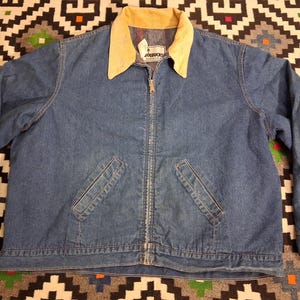 Könnte beinhalten: Eine blaue Jeansjacke mit beigem Cordkragen, durchgehendem Frontreißverschluss und zwei schrägen Leistentaschen an der Taille. Das Innenetikett zeigt den Text Roebucks, mit langen Ärmeln und geknöpften Manschetten.