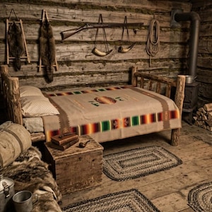 Puede incluir: Un dormitorio rústico de cabaña de troncos con una estructura de cama de madera y una manta color canela con un borde geométrico colorido. La habitación incluye un cofre de madera, un saco de dormir de lona enrollado, pieles, un rifle y chimenea.