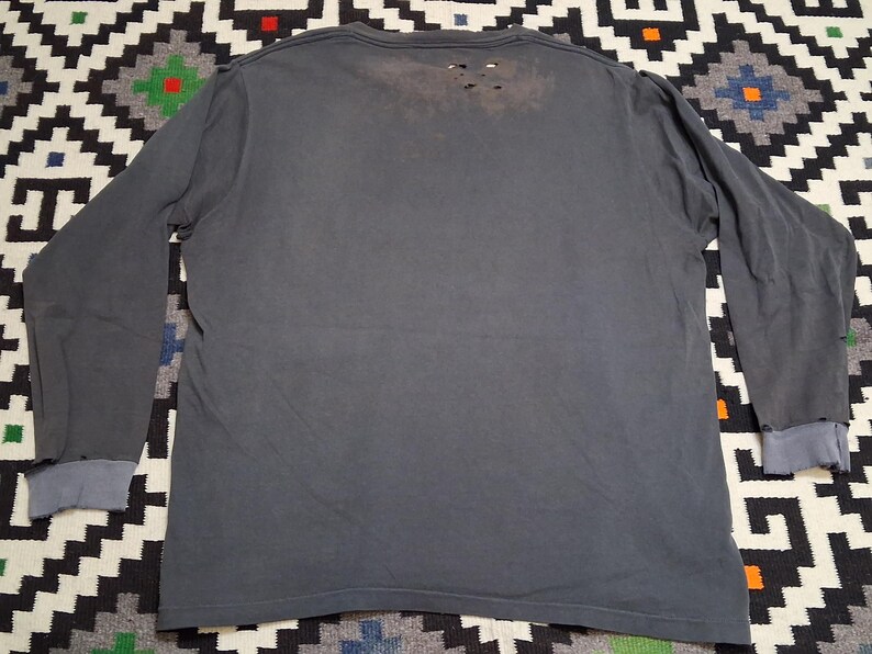 Puede incluir: Una camiseta de manga larga gris carb&oacute;n con detalles desgastados. La camiseta tiene cuello redondo y pu&ntilde;os en un gris m&aacute;s claro. La parte trasera de la camiseta tiene varios agujeros peque&ntilde;os cerca del cuello, d&aacute;ndole un aspecto usado.