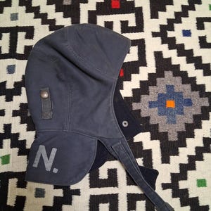 May include: A dark blue winter hat with a hood and ear flaps. The hat has a strap with a button closure and the letter "N." printed on the side in a lighter color. The hat is made of a durable fabric.