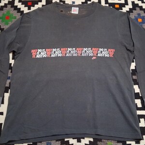 Puede incluir: Camiseta de manga larga gris oscuro con el texto "JUST DO IT" en rojo y blanco repetido en el pecho. Un peque&ntilde;o logotipo de Nike es visible. La camiseta tiene cuello redondo y pu&ntilde;os doblados.