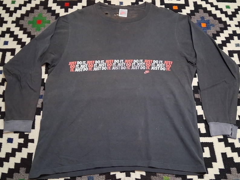Puede incluir: Camiseta de manga larga gris oscuro con el texto "JUST DO IT" en rojo y blanco repetido en el pecho. Un peque&ntilde;o logotipo de Nike es visible. La camiseta tiene cuello redondo y pu&ntilde;os doblados.
