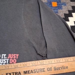 Puede incluir: Camiseta de manga larga gris oscuro con el texto "DO IT. JUST DO" en rojo y blanco. La camiseta es parcialmente visible, con un desgarro en la manga. Una regla de madera con texto est&aacute; en primer plano.