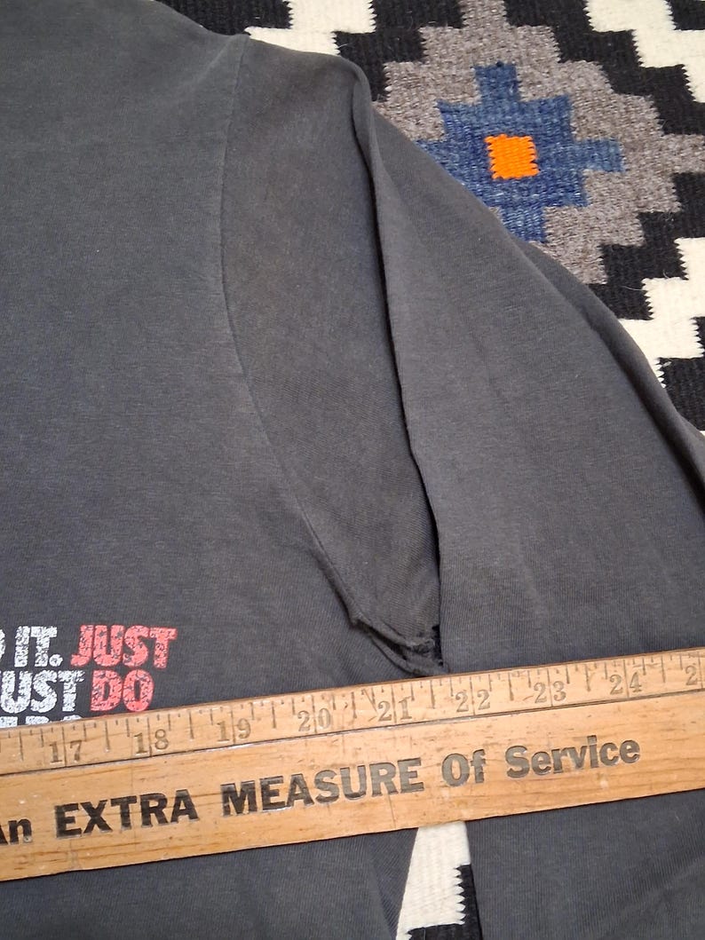 Puede incluir: Camiseta de manga larga gris oscuro con el texto "DO IT. JUST DO" en rojo y blanco. La camiseta es parcialmente visible, con un desgarro en la manga. Una regla de madera con texto est&aacute; en primer plano.