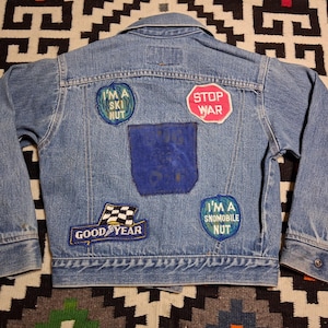 May include: A light blue denim jacket featuring a collar, long sleeves with buttoned cuffs, and several patches on the back. The patches include text reading I'M A SKI NUT, STOP WAR, GOOD YEAR, and I'M A SNOMOBILE NUT, alongside a dark blue fabric square.
