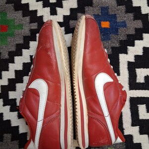 May include: Pair of red sneakers with white accents, including the iconic swoosh logo on the sides. The shoes have white laces and soles. The sneakers are displayed on a patterned rug.