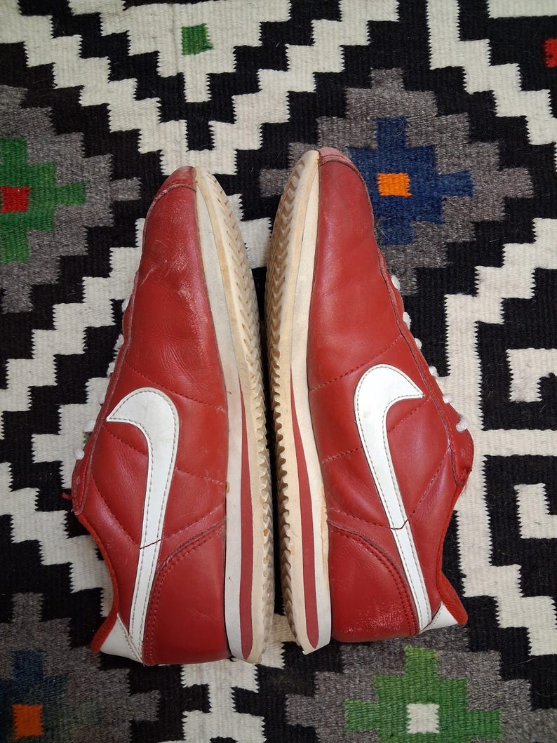 May include: Pair of red sneakers with white accents, including the iconic swoosh logo on the sides. The shoes have white laces and soles. The sneakers are displayed on a patterned rug.