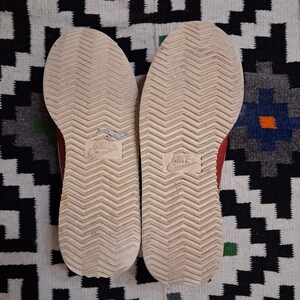 May include: Pair of red and white sneakers, soles facing up, showing the tread pattern and the Nike logo. The shoes are worn, with some visible wear and tear. The background is a patterned rug with black, white, and blue geometric shapes.