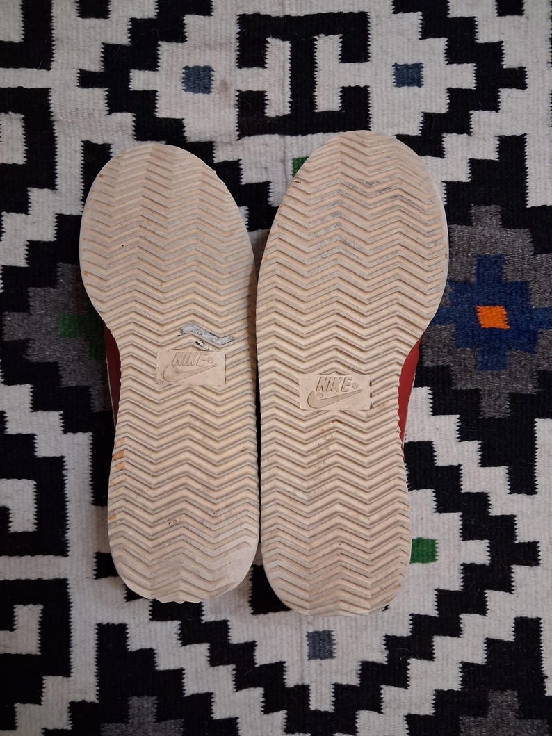 May include: Pair of red and white sneakers, soles facing up, showing the tread pattern and the Nike logo. The shoes are worn, with some visible wear and tear. The background is a patterned rug with black, white, and blue geometric shapes.