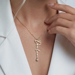 May include: A gold-colored necklace with the name "Destiny" in cursive script. The pendant hangs vertically and features a small, round, clear gemstone at the bottom. The necklace is worn over a white blazer.