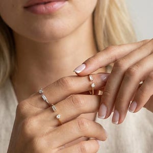 May include: A collection of delicate gold rings adorned with small, sparkling gemstones. The rings feature various designs, including baguette and round-cut stones. The image showcases the rings on a person's fingers, highlighting their elegant and minimalist style.