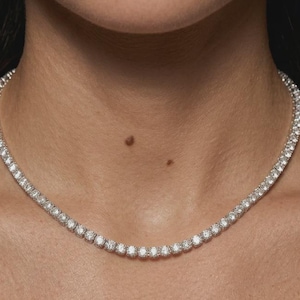 May include: A silver-toned tennis necklace featuring a continuous row of round, clear, faceted stones set in a metallic frame. The necklace rests closely around the neck, showcasing a uniform design of sparkling, circular gems linked together.