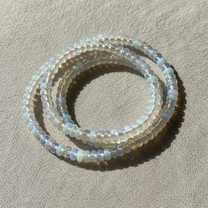 May include: Three strands of iridescent beaded bracelets, coiled on a neutral-toned fabric. The beads have a subtle, opalescent sheen, reflecting a range of colors. The bracelets are a delicate accessory.