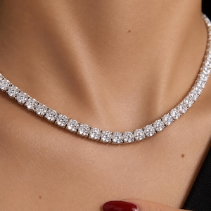 May include: A silver-toned tennis necklace featuring a continuous row of round, brilliant-cut clear stones set in a metallic frame. The necklace rests against the neck, showcasing a uniform arrangement of sparkling, faceted stones throughout the design.