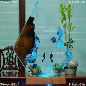 Puede incluir: Una escultura decorativa de resina y madera con una losa de madera oscura integrada en resina azul translúcida. La pieza representa una escena submarina con dos siluetas negras de buceadores, una tortuga marina y detalles de coral.