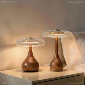 May include: Two table lamps with smooth, dark wood bases and wide, clear glass disc shades. Each lamp features a central metallic accent connecting the base to the glass top, emitting a soft, warm glow from the internal light source.