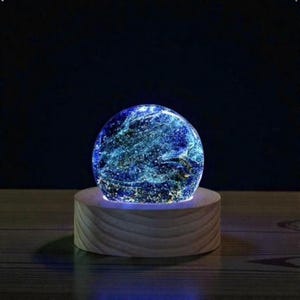 Puede incluir: Una esfera azul brillante con un diseño similar a una galaxia, colocada sobre una base de madera. La esfera emite una luz suave y ambiental, creando un efecto celestial. La base de madera es de color marrón claro.
