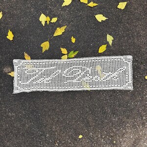 May include: A rectangular white crochet doily featuring an intricate openwork grid pattern. The center of the piece displays the cursive script text "La Vie Est Belle" woven into the thread work, creating a delicate and detailed textile design.