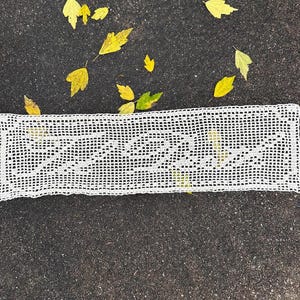 May include: An off-white, crocheted table runner with a rectangular shape and an openwork design. It rests on dark asphalt, surrounded by yellow autumn leaves. The runner's detailed pattern is clearly visible, showcasing the craftsmanship.