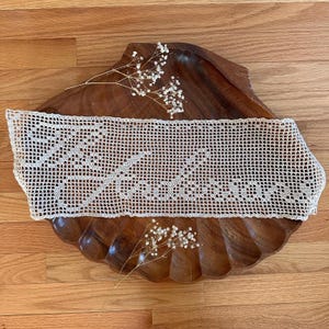 May include: Cream-colored, open-weave crochet doily with a geometric pattern, resting on a dark wooden, shell-shaped bowl. The doily is rectangular and features a repeating design. Small white flowers are placed near the doily.