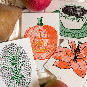 Könnte beinhalten: Vier kleine rechteckige Kunstdrucke mit Siebdruck-Illustrationen einer grünen Gottesanbeterin, eines orangefarbenen Kürbisses, einer grünen Teetasse und einer orangefarbenen Lilie. Jeder Druck enthält handschriftliche Beschriftungen und Nummerierungen.