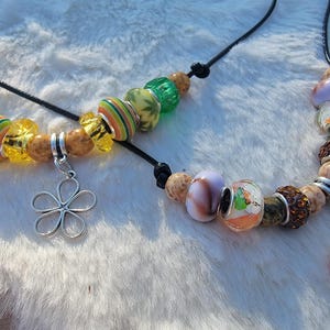 May include: Two beaded necklaces with a variety of colorful beads. One necklace features a silver flower charm, while the other has a teardrop-shaped charm. The beads are in shades of green, yellow, orange, brown, and purple.