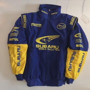 Könnte beinhalten: Eine blaue und gelbe Jacke des Subaru World Rally Teams. Die Jacke zeigt das Subaru-Logo, Sponsorenlogos und Text. Die Ärmel sind gelb und der Körper ist blau. Die Jacke hat einen durchgehenden Reißverschluss und einen Stehkragen.