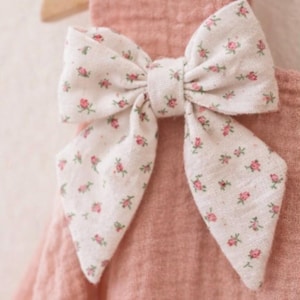 May include: A pink fabric item with a large bow. The bow is white with a pattern of small pink flowers and green leaves. The item is hanging on a wooden hanger.