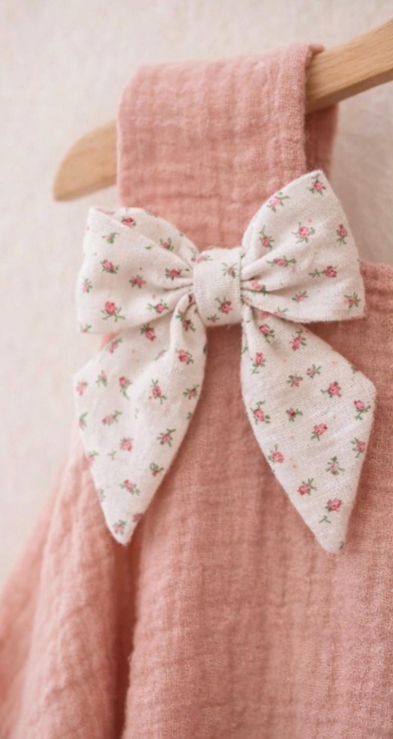 May include: A pink fabric item with a large bow. The bow is white with a pattern of small pink flowers and green leaves. The item is hanging on a wooden hanger.