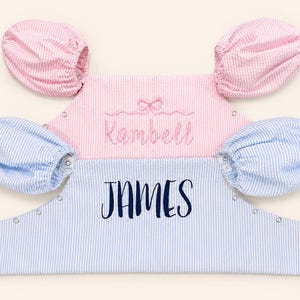 May include: Two seersucker rompers with puff sleeves and metal snap closures. The top romper is pink with white stripes, featuring an embroidered pink bow and the name Kambell. The bottom romper is blue with white stripes and the name JAMES.