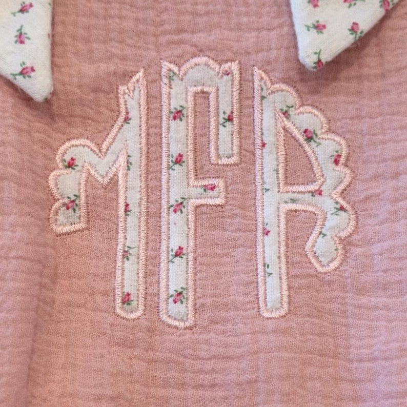 May include: Close-up of a pink fabric with the embroidered letters "MFH" in a floral pattern. The letters are outlined in pink and filled with a white fabric adorned with small pink flowers. The background is a textured pink fabric.