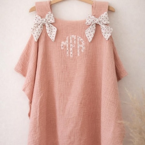 May include: A light pink, muslin fabric cover-up with a monogram and floral bow accents. The cover-up is hanging on a wooden hanger. The bows are white with a small pink floral pattern.