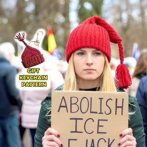 May include: A red knitted hat with a long tassel and a matching keychain. The keychain is a miniature version of the hat. The image also includes the text "ABOLISH ICE" on a cardboard sign.